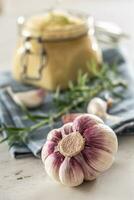 einheimisch Knoblauch Glühbirnen im Vorderseite von ein Glas Krug voll von frisch Knoblauch Einfügen gelegt auf Küche Stoff mit Rosmarin foto