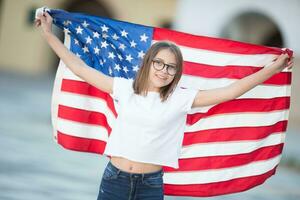 glücklich jung amerikanisch Schule Mädchen halten und winken im das Stadt mit USA Flagge foto