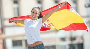 glücklich lächelnd ziemlich Mädchen hält ein Flagge von Spanien hinter ihr feiern auf ein Straße foto