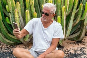 gut aussehend Mitte alt Mann mit Smartphone im Vorderseite von ein groß Kaktus im ein exotisch Strand Resort foto