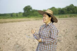 asiatisch Frau Farmer ist beim Landwirtschaft Land, tragen Hut, Plaid Shirt, hält und schreiben auf Papier Zwischenablage. Konzept , Forschung und analysieren Boden und Umgebung zu verbessern und lösen Landwirtschaft Probleme. foto
