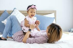 asiatisch Mutter ist spielen mit ihr ziemlich lächelnd Baby Tochter während Ausgaben Qualität Zeit im das Bett zum Familie Glück und Erziehung Konzept foto