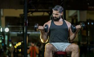 kaukasisch Bart muskulös Sport Mann ist trainieren Gewicht Ausbildung auf doppelt Hanteln zum Bizeps und Trizeps Muskel Innerhalb Fitnessstudio mit dunkel Hintergrund zum ausüben und trainieren Konzept foto