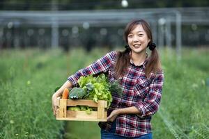 asiatisch Frau Farmer ist Tragen hölzern Tablett voll von frisch wählen Bio Gemüse im ihr Garten zum Ernte Jahreszeit und gesund Diät Essen Konzept foto