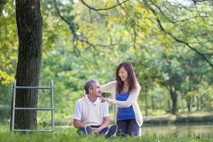 asiatisch Senior Vater und Tochter Lachen während glücklich Sitzung zusammen unter das groß Baum im das Park haben Licht Massage während Sommer- zum mental Gesundheit und Langlebigkeit Konzept foto