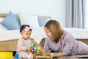 asiatisch Mutter ist spielen mit ihr ziemlich lächelnd Baby Tochter mit hölzern Spielzeug Block während Ausgaben Qualität Zeit im das Bett zum Familie Glück und Erziehung Konzept foto