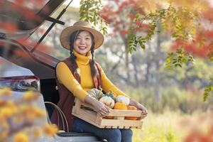 glücklich asiatisch Farmer Mädchen Tragen produzieren Ernte mit einheimisch Bio Apfel, quetschen und Kürbis Sitzung auf das Auto Kofferraum beim lokal Bauernhof Markt während Herbst Jahreszeit zum Landwirtschaft Produkt foto