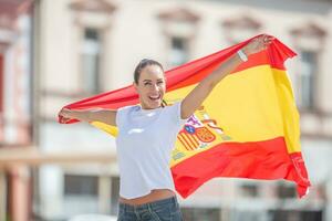 glücklich lächelnd ziemlich Mädchen hält ein Flagge von Spanien hinter ihr feiern auf ein Straße foto