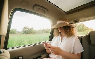 heiter positiv lockig jung Frau im beiläufig tragen mit Hut Sitzung im Automobil Rücksitz mit befestigt Sicherheitsgurt und mit Telefon. foto