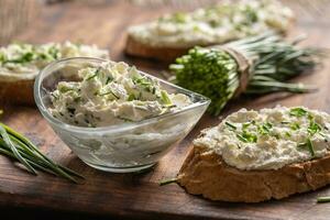 ein Schüssel von hausgemacht Sahne Käse Verbreitung mit gehackt Schnittlauch umgeben durch Brot Scheiben mit Verbreitung und ein Bündel von frisch Schnitt Schnittlauch foto