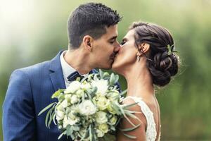 Jungvermählten Kuss draußen während halten ein Hochzeit Strauß von Weiß Blumen im Vorderseite von Sie foto