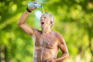 alt Mann mit Nein Hemd gießt Wasser von ein Flasche Über seine Kopf und Gesicht draußen auf ein heiß Sommer- Tag foto