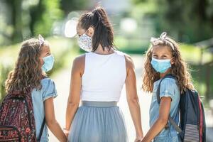 ein Mutter und ihr Zwilling Tochter verwenden ein schützend Maske wann Rückkehr zu Schule während das covid-19 Quarantäne foto