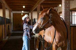 jung Mädchen im Cowboy Hut Kämme das eingestallt Pferd Das sieht aus zurück beim ihr drinnen foto