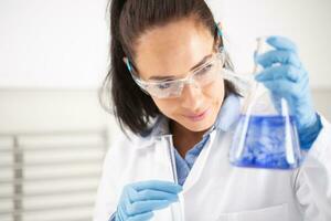 weiblich Chemiker tragen schützend Brille lächelt wann halten ein Reagenzglas, Mischen Blau Flüssigkeit Innerhalb das konisch Flasche foto