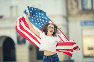 glücklich jung amerikanisch Schule Mädchen halten und winken im das Stadt mit USA Flagge foto