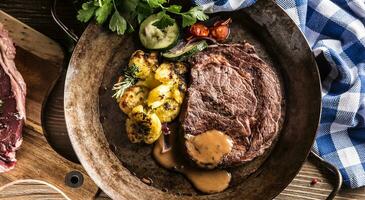 saftig Rindfleisch Rippe Auge Steak geröstet Kartoffeln mit Soße gegrillt Gemüse Salz- Pfeffer und Kräuter im Jahrgang schwenken foto