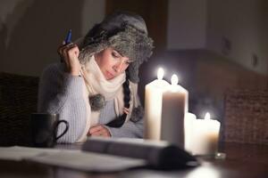 Frau sitzt im Winter Hut beim hoem tun Papierkram unter Beleuchtung von Kerzen wie Russland Schnitt Gas liefert und Europa leidet von Energie Krise foto
