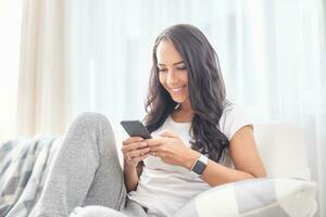 lächelnd jung weiblich Sitzung auf ein Couch im ein Leben Zimmer lächelnd in das Telefon Ausgaben Zeit online während ein Sperrung foto