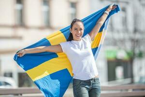 lächelnd weiblich Ventilator hält Schwedisch Flagge auf ein Straße foto