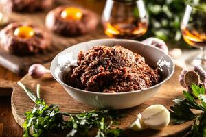 Weiß Schüssel gefüllt mit roh Fleisch bereit zu bereiten köstlich Steak Tartar mit Bohnenkraut Ei Eigelb. schon bereit Tataren im das Hintergrund mit Rum und Kräuter wie ein Dekoration foto