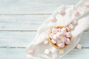 oben Aussicht von ein hölzern Schüssel voll von Rosa und Weiß Marshmallows mit etwas verstreut um auf ein Weiß Tabelle Stoff und Weiß hölzern Oberfläche foto