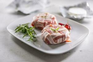 Camembert oder Brie Käse eingewickelt im Speck mit frisch Rosmarin und Chili auf ein Weiß Teller foto