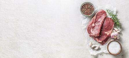 zwei Rippe Auge Steak Salz- Gewürze Rosmarin und Knoblauch oben von Sicht. Rindfleisch Fleisch auf Weiß Beton Tabelle foto