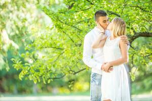 jung Paar küssen unter das Grün Baum mit Sommer- Natur Hintergrund. gekleidet im Weiß Kleid, Weiß Hemd und Jeans, halten Hände foto