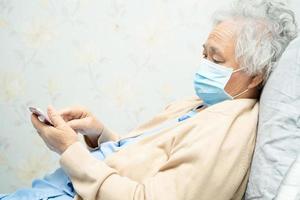 asiatische ältere Patientin, die eine Gesichtsmaske trägt und ein Mobiltelefon im Krankenhaus hält, um die Sicherheitsinfektion zu schützen und das neuartige Coronavirus Covid-19-Virus abzutöten.