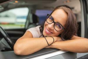 lächelnd weiblich Treiber tragen Brille lehnt sich durch das öffnen Tür Fenster Sitzung auf Fahrer Sitz foto