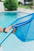 Hände halten ein Skimmer mit Blau Schwimmbad im das Hintergrund, ein Mann Reinigung Schwimmbad mit Blatt Skimmer. Mann Reinigung das Schwimmbad mit das Abschäumer, Person mit Skimmer Reinigung Schwimmbad foto