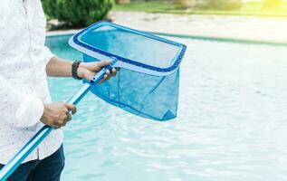 Hände halten ein Skimmer mit Blau Schwimmbad im das Hintergrund. ein Mann Reinigung Schwimmbad mit Blatt Skimmer. Mann Reinigung das Schwimmbad mit das Abschäumer, Person mit Skimmer Reinigung Schwimmbad foto