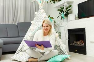 blond Frau Sitzung auf ein Fußboden mit ein Buch im das Weiß Zimmer in der Nähe von Wigwam foto