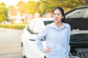 Junge Frau, die in der Nähe eines kaputten Autos mit aufgeklappter Motorhaube steht und Probleme mit ihrem Fahrzeug hat foto