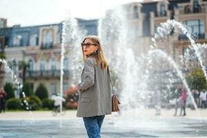 schön weiblich Frau tragen passen gehen in der Nähe von Stadt Brunnen. städtisch Porträt von jung elegant Geschäft Frau mit Brille und beiläufig Kleidung. Hintergrund von schön architektonisch Gebäude. foto