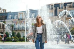 Mädchen beim Brunnen sieht aus direkt beim Kamera. Mädchen Gehen um Stadt gegen Hintergrund von schön architektonisch Gebäude. streng Geschäftsfrau tragen Jacke und Jeans. verschwommen Hintergrund. foto