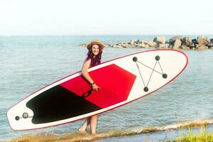 sup einsteigen. ein Frau im ein Hut Spaziergänge mit ein sup Tafel. das Konzept von aktiv Lebensstil und Surfen. foto