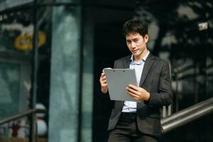 zuversichtlich asiatisch Mann mit ein Lächeln Stehen halten Notizblock und Tablette beim das modern Büro. foto