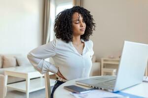 zurück Schmerzen Schlecht Haltung Frau Sitzung im Büro. jung afro Frau Leiden von zurück Schmerzen während Arbeiten auf Laptop beim Zuhause foto