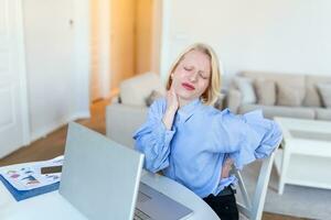 Porträt von jung betont Frau Sitzung beim Zuhause Büro Schreibtisch im Vorderseite von Laptop, berühren Schmerz zurück mit schmerzte Ausdruck, Leiden von Rückenschmerzen nach Arbeiten auf Laptop foto