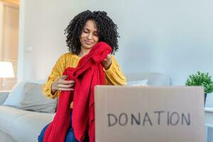 glücklich afrikanisch jung Frau sitzen auf Couch stecken Kleider im Spende Box beim heim, Pflege biracial weiblich Freiwillige stellen bekleidung im Karton Paket, spenden zu Bedürftige Menschen, Wiederverwendung, recyceln Konzept foto