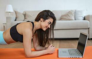 Porträt von jung attraktiv Frau tun Übungen während Aufpassen online Lektionen auf ihr Laptop. gesund Lebensstil und Sport Konzept. Schuss von Fitness Frau Sitzung auf Übung Matte foto