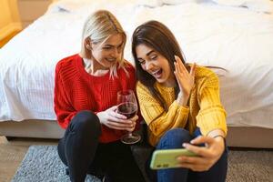 freunde mit Wein auf das Fußboden beim heim. glücklich lächelnd jung Frauen freunde suchen beim Fotos auf Handy, Mobiltelefon Telefon. Mädchen havin Spaß. zwei weiblich freunde entspannend beim Zuhause mit Glas von Wein reden zusammen