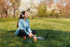Foto einer jungen freiberuflichen Frau, die im Stadtgras im Park sitzt und Musik vor Laptop und Kaffeetasse hört?