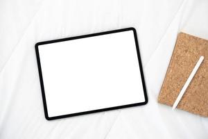 Draufsicht auf Tablet mit weißem Bildschirm liegt auf einem Bett mit Notebook und digitalem Stift. foto