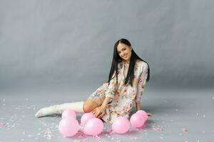 ein ziemlich jung Frau sitzt in der Nähe von Rosa Luftballons und Konfetti auf ein grau Studio Hintergrund. foto