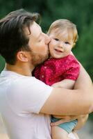 ein jung Vater Küsse seine Sohn auf das Wange während Gehen im das Park im Sommer- foto