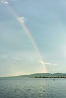 mehrfarbig schön Regenbogen Über das Fluss Bucht im das Himmel mit Wolken. natürlich natürlich Phänomen. Vertikale Aussicht foto