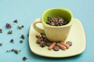 ein Tasse mit getrocknet Oregano und Kräuter- medizinisch Kapseln und Tabletten auf ein Untertasse auf ein Grün Hintergrund. Alternative Medizin foto